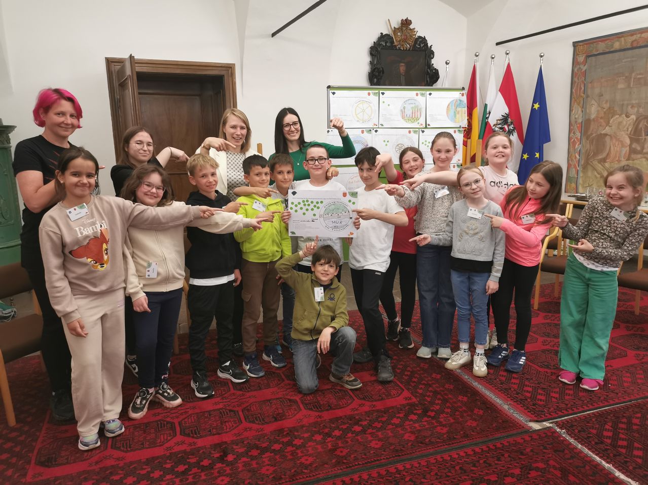 A group of children and adults stand and crouch on a red carpet, holding a piece of paper and pointing at it. Behind them are flags and a wall with a poster.