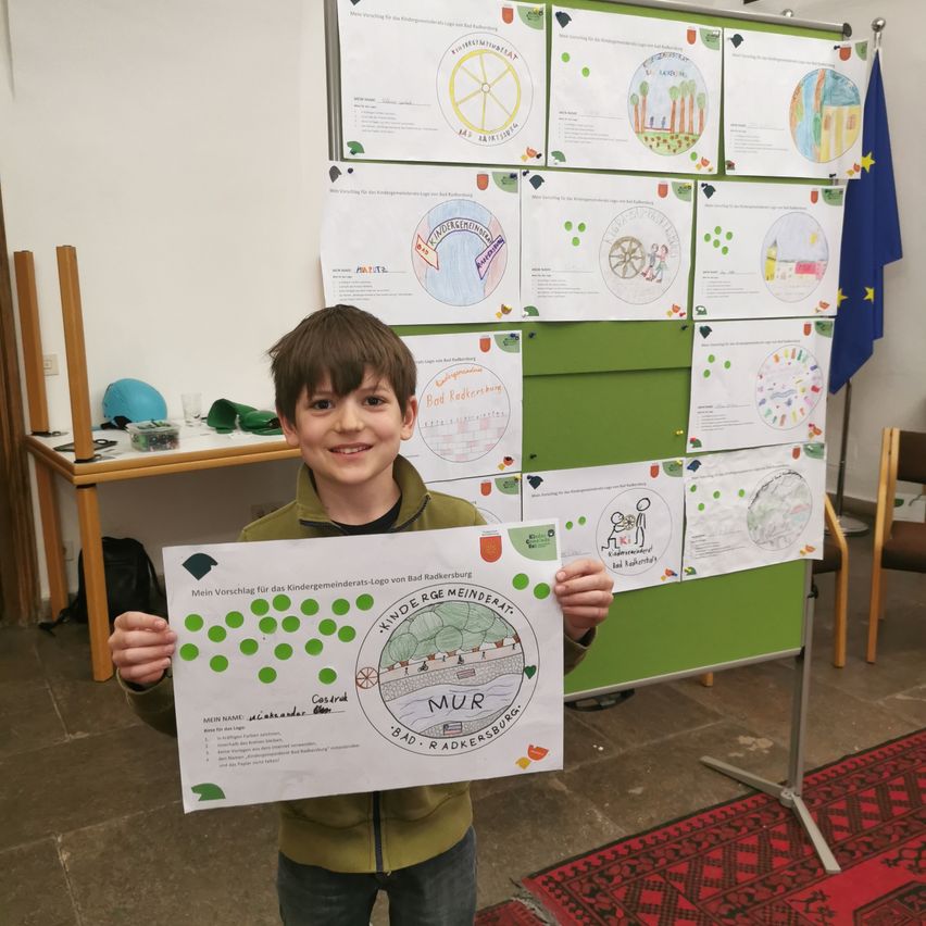 A young boy holding a poster for the Kindergemeinderat of Bad Radkersburg, surrounded by other posters and a European flag.