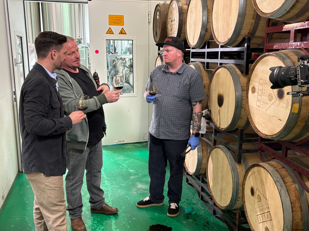 Three men are in a room with barrels and glass cups of wine. One is holding a drill.