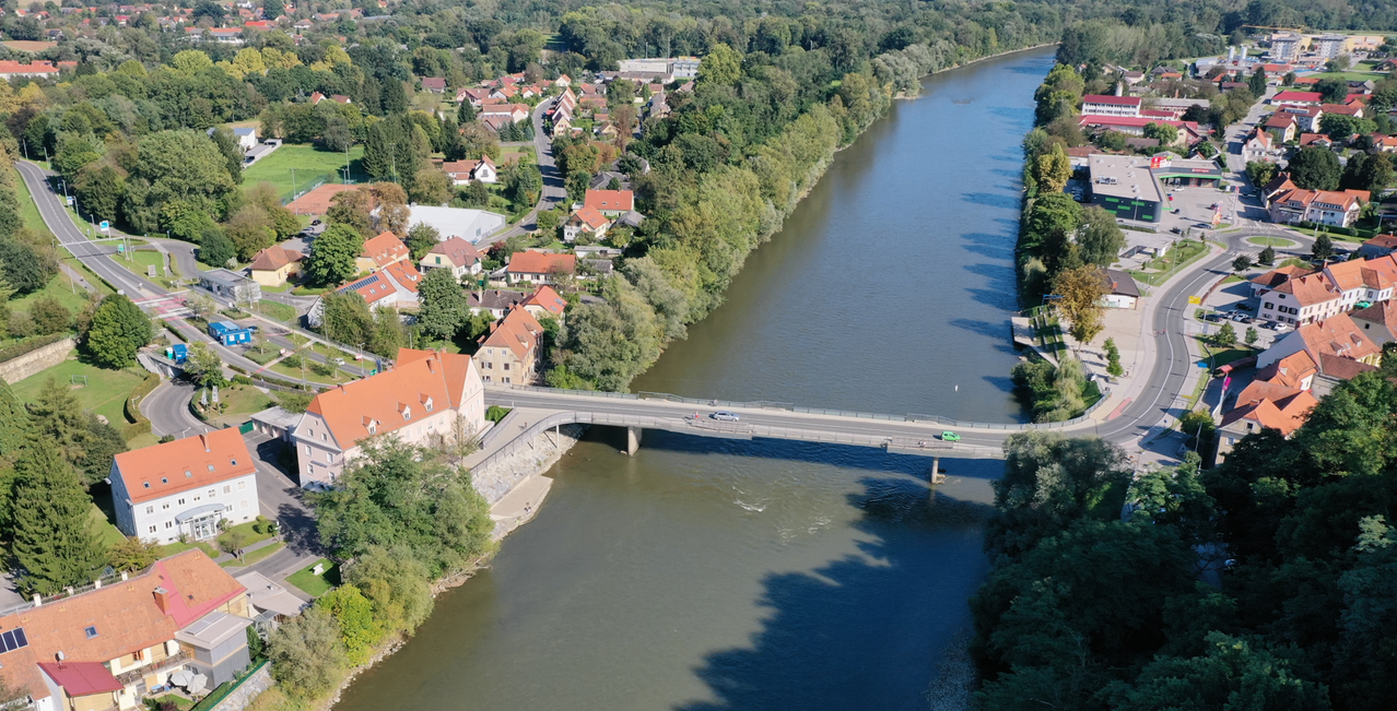 An aerial view of a river with a bridge over it, houses, and trees along the banks. Cars are driving on the bridge.