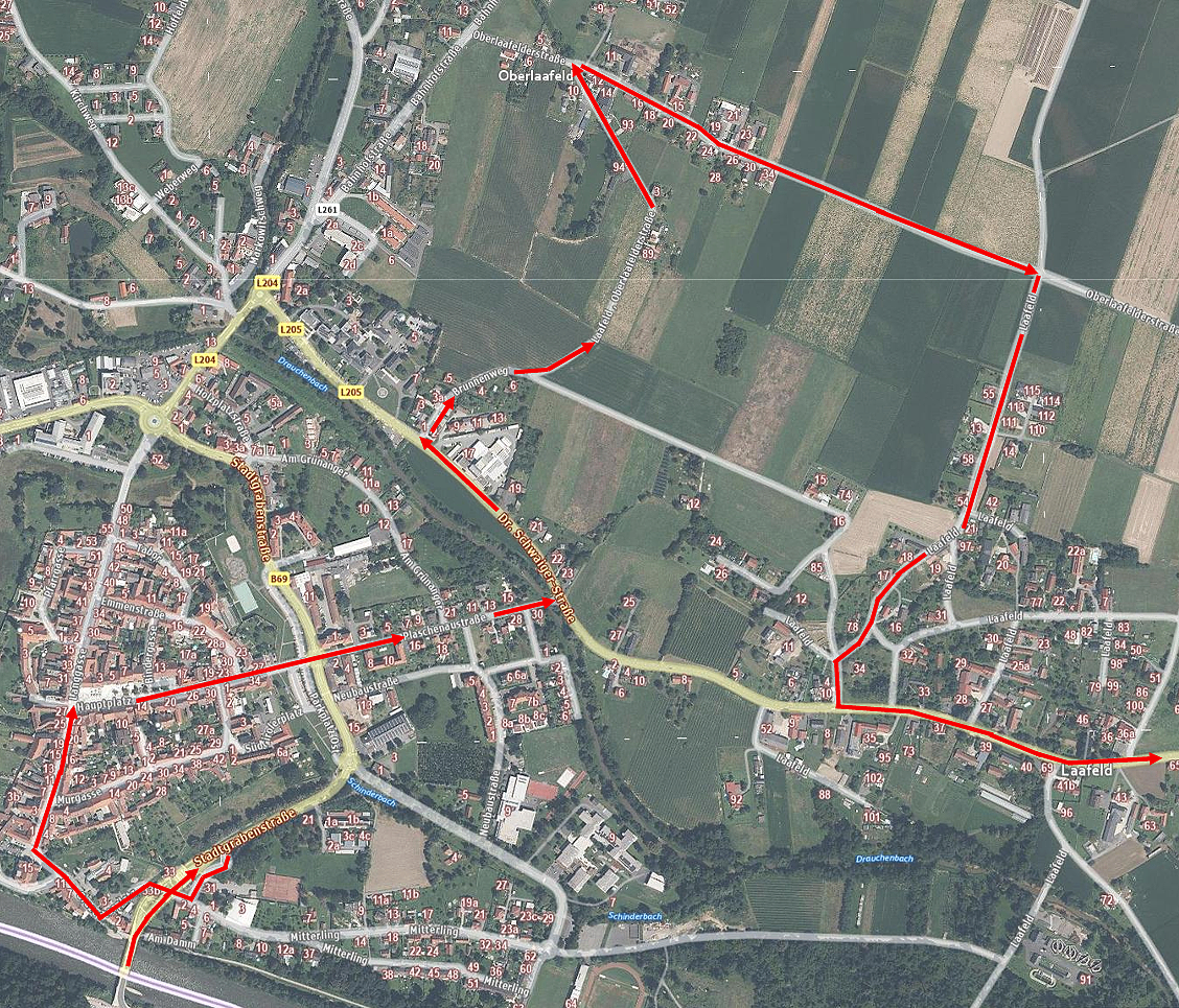 Luftaufnahme einer Stadt mit in Rot markierten Straßen und Wegen. Die Straßen sind nummeriert und beschriftet. Grüne Felder und Bäume sind sichtbar.