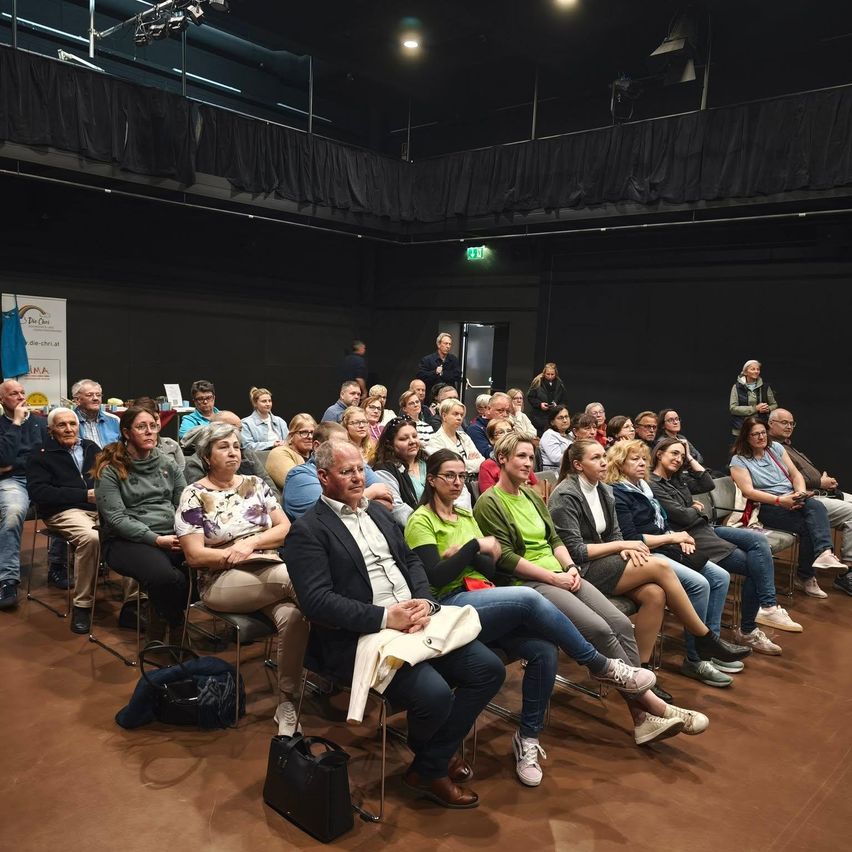 Eine Gruppe verschiedener Personen sitzt aufmerksam in einem Theater und hört einem Redner zu. Einige tragen Brillen, Sneakers und Mäntel. Stühle, Taschen und eine Bühne mit einem Banner sind sichtbar.