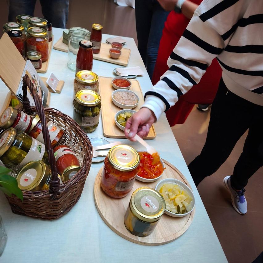 Eine Person mit einem gestreiften Pullover löffelt Essen aus einem Glas auf einem Tisch. Der Tisch hat viele Gläser und Schalen mit Essen. Links steht ein Korb mit weiteren Gläsern.