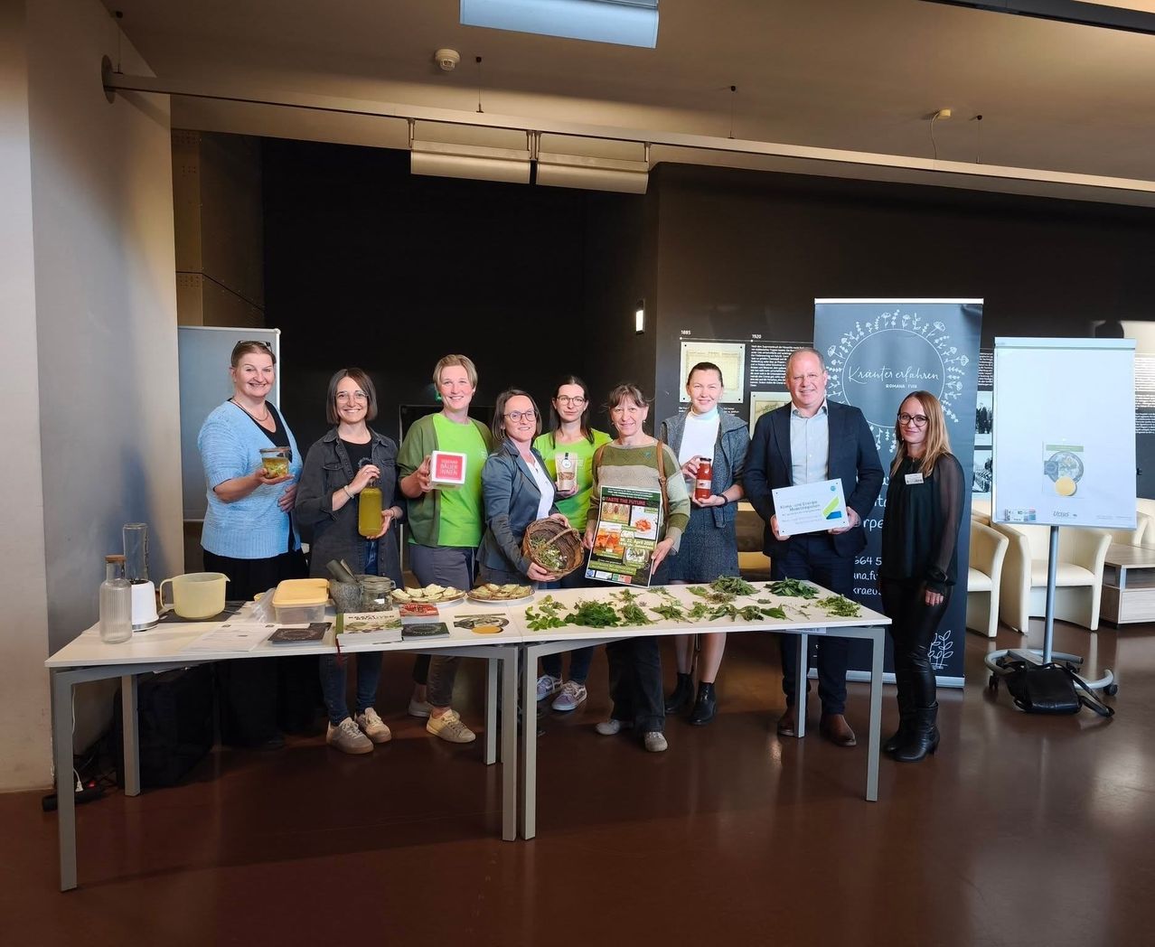 Eine Gruppe von Menschen steht um einen Tisch herum, der mit verschiedenen Lebensmitteln und Flaschen gefüllt ist. Einige halten Auszeichnungen und Bücher, während andere Gläser mit Getränken haben. Sie scheinen ein Ereignis zu feiern.
