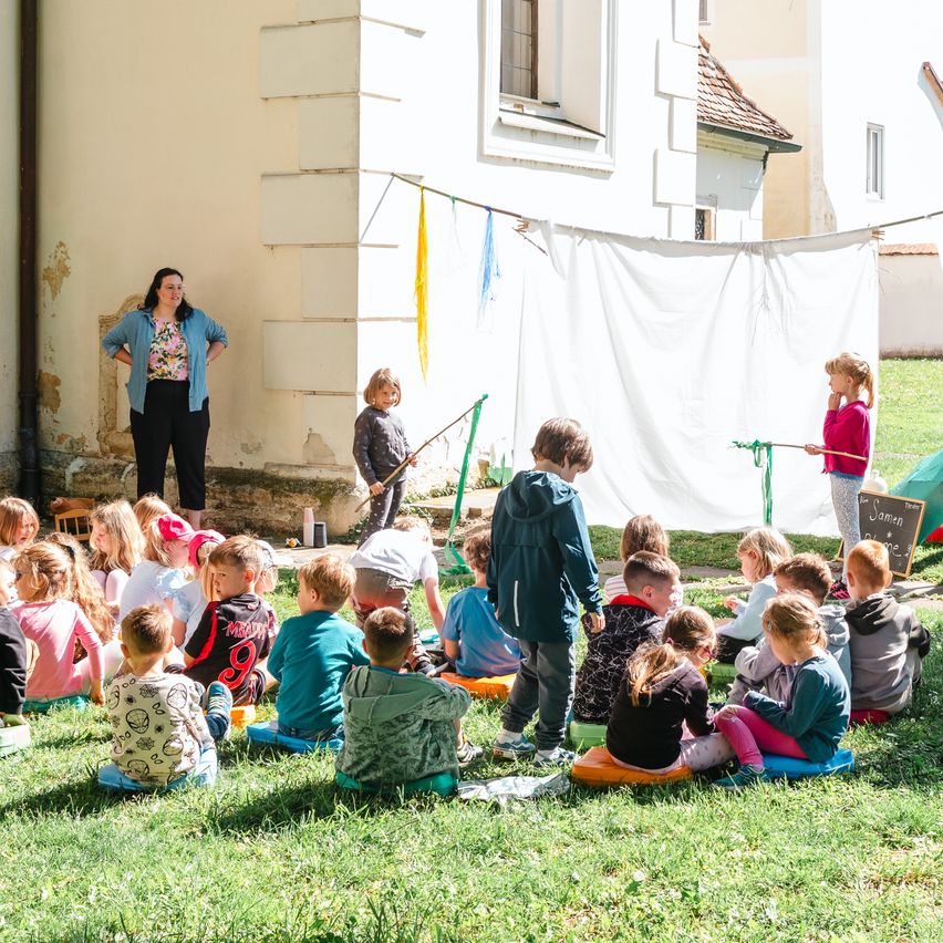 Eine Gruppe von Kindern sitzt auf dem Rasen vor einem Gebäude, mit einer Frau in der Nähe. Einige halten Stöcke und eine Tafel ist im Hintergrund.