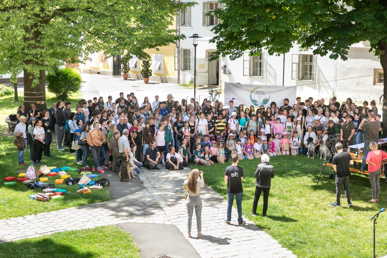 Eine große Gruppe von Menschen ist auf einem grünen Rasen versammelt. Sie stehen und sitzen vor einem Gebäude mit vielen Fenstern. Einige Menschen machen Fotos.