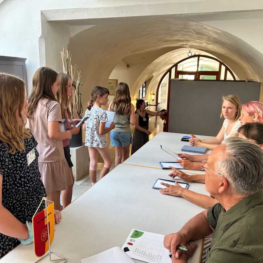 A group of young women and men are gathered around a table with papers and books, likely participating in a workshop or meeting in a historic building.