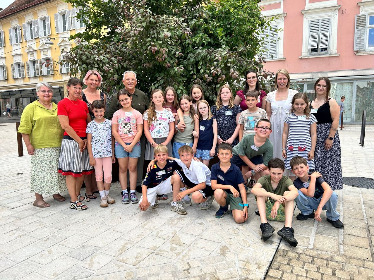 A group of adults and children poses for a photo in front of a tree in a city.