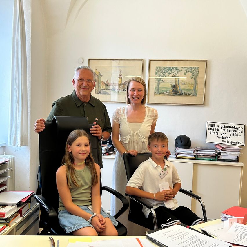 An older man and woman, along with two children, sit in office chairs in a room with a desk. The man and woman stand behind the children, and the room has paintings, a sign, and a desk with papers.