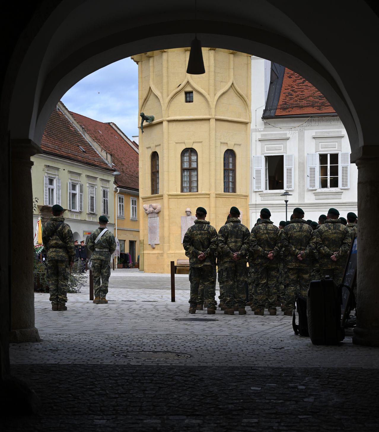Eine Gruppe von Soldaten steht in Formation auf einer Kopfsteinpflasterstraße, mit einem gelben Gebäude und einer Statue dahinter.