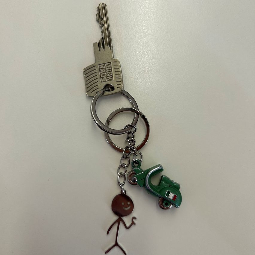 Ein silberner Schlüsselanhänger mit einem Schlüssel, einem grünen Roller-Anhänger und einer braunen Strichfigur auf weißem Hintergrund.