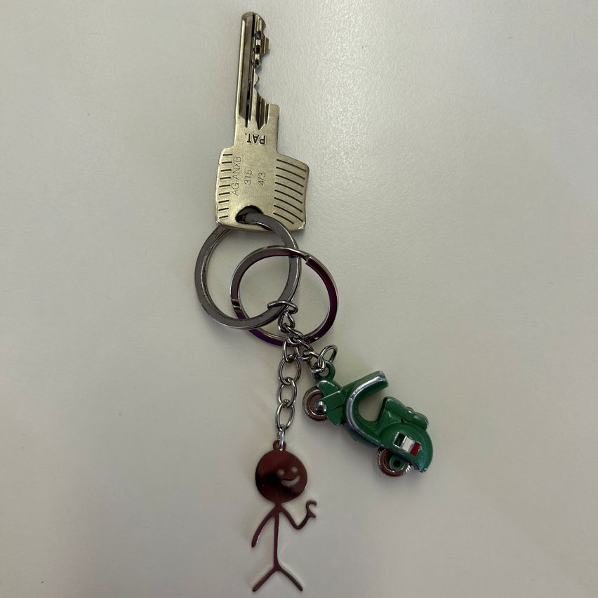 Ein silberner Schlüsselanhänger zeigt einen Schlüssel mit 'PAD' darauf. An den Schlüssel ist eine Kette mit einem kleinen grünen Roller-Anhänger und einer braunen Figur befestigt.