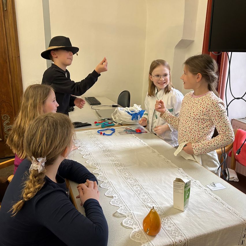 Mehrere Kinder in einem Klassenzimmer, ein Kind in Hut und Mikrofon gestikuliert, während ein anderes Kind ein Blatt Papier hält. Andere Kinder sitzen an einem Tisch mit Tischdecke und verschiedenen Gegenständen.