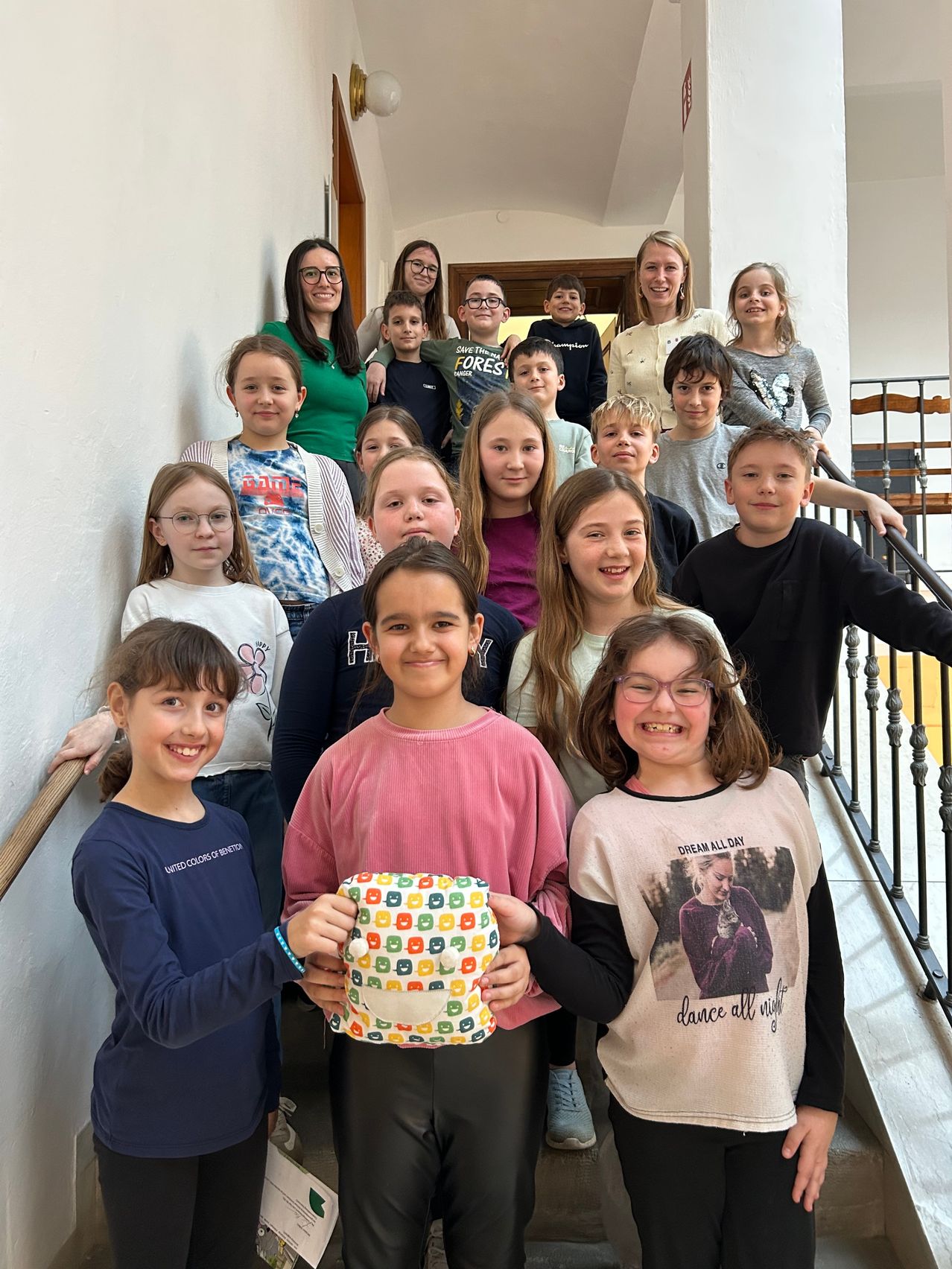 Eine Gruppe von Kindern und einigen Erwachsenen steht auf der Treppe und lächelt für ein Foto. Die Kinder tragen Freizeitkleidung, einige mit Brillen. Ein Kind hält ein farbenfrohes Objekt.