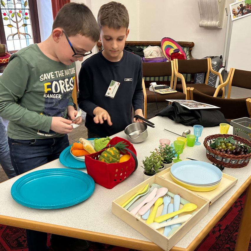 Zwei Jungen stehen bei einem Tisch, der für eine Mahlzeit mit Obst, Besteck und Tellern gedeckt ist. Ein Junge hält einen Löffel, während der andere in den Obstkorb schaut.