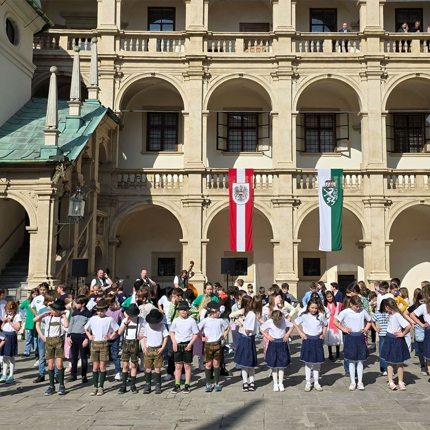 Kinder in traditioneller Kleidung stehen auf dem Innenhof eines historischen Gebäudes. Flaggen hängen vom Balkon. Einige Kinder spielen Musikinstrumente.