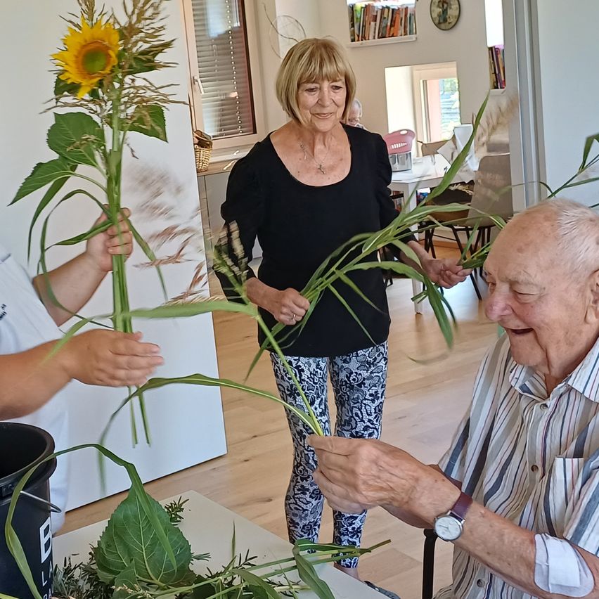 Ein älterer Mann und eine ältere Frau ordnen Sonnenblumen und Blätter auf einem Tisch an, während ein Kind im Hintergrund eine Sonnenblume hält.