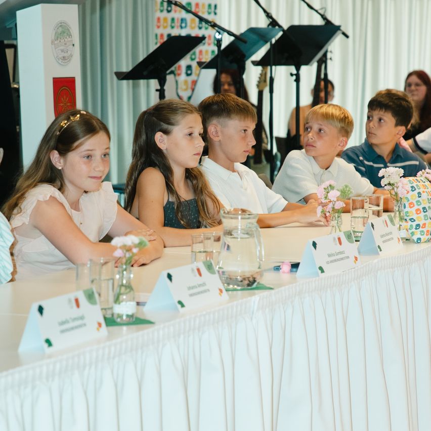 Fünf Kinder sitzen aufmerksam an einem Tisch mit Namenskarten und Blumen, wahrscheinlich in einer Kirche. Musikinstrumente und Erwachsene sind im Hintergrund.