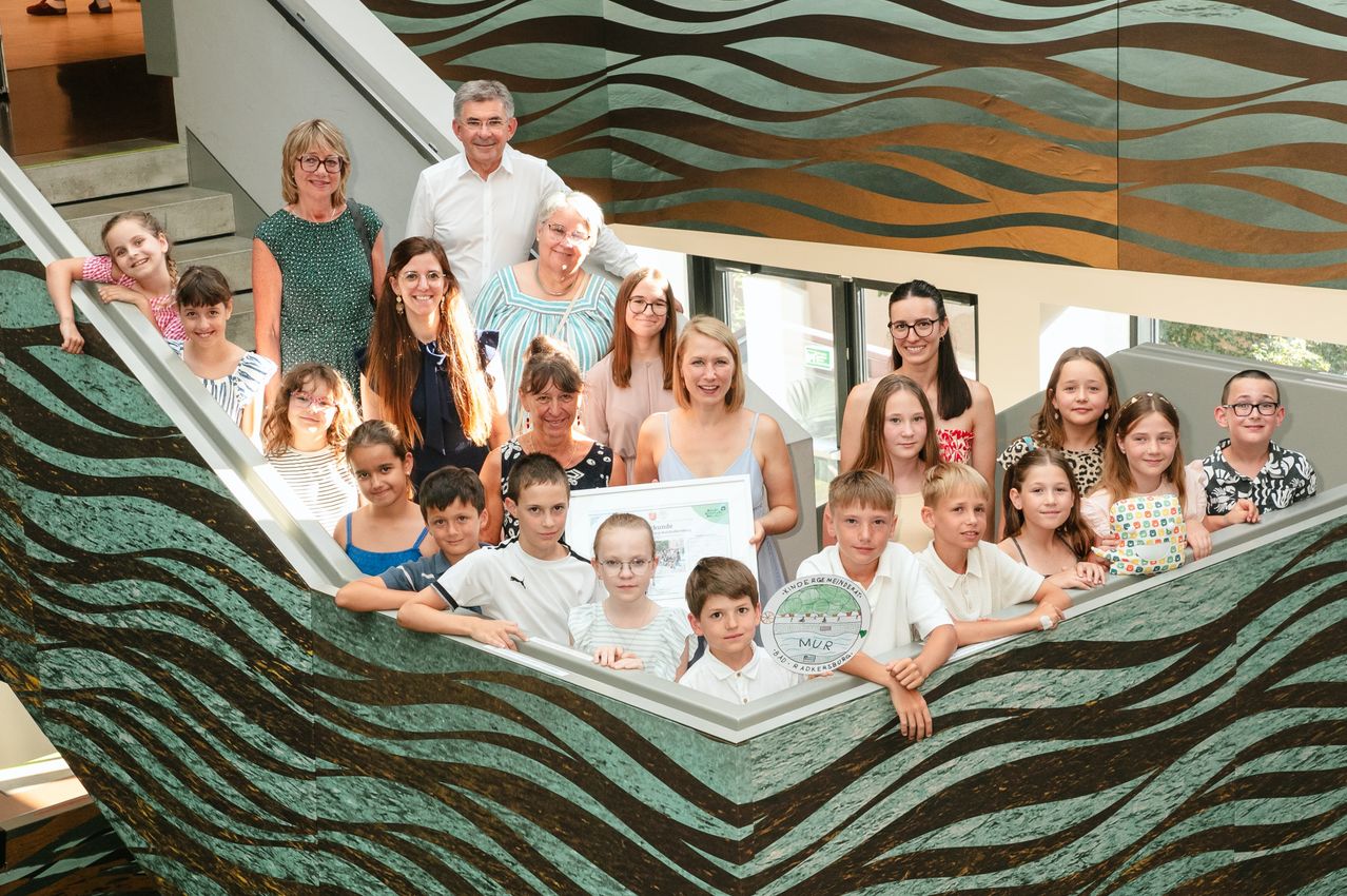 Eine Gruppe von Menschen posiert für ein Foto auf einer Treppe. Sie halten eine Plakette und lächeln für die Kamera. Ein Hintergrund mit Wellenmustern ist sichtbar.