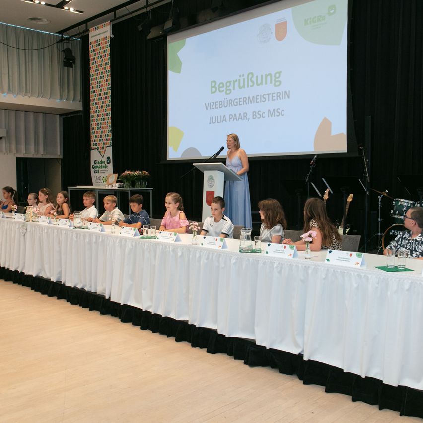 Eine Frau spricht an einem Podium vor einem Bildschirm, der 'Begrüßung' und Namen anzeigt. Kinder sitzen an einem Tisch mit Blumen und Namensschildern. Musikinstrumente sind vorhanden.