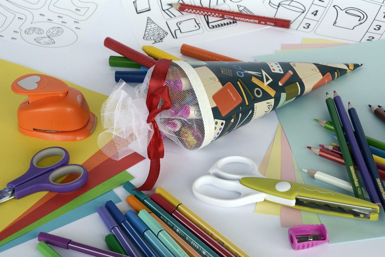 Eine Auswahl an bunten Kunstmaterialien auf einem Tisch, einschließlich einer kegelförmigen Tasche mit einer Schleife, Scheren und Bleistiften.
