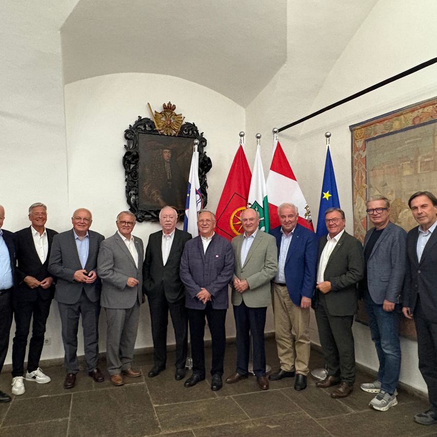 Eine Gruppe von Männern in Anzügen steht vor einer Wand mit Flaggen und einem Gemälde.