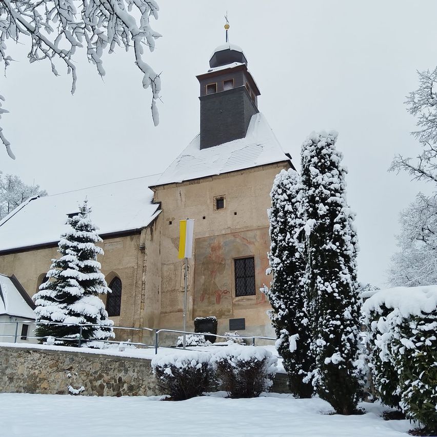 Eine Kirche mit schneebedecktem Dach und einem Turm, umgeben von schneebedeckten Bäumen, Büschen und einem Fahnenmast.