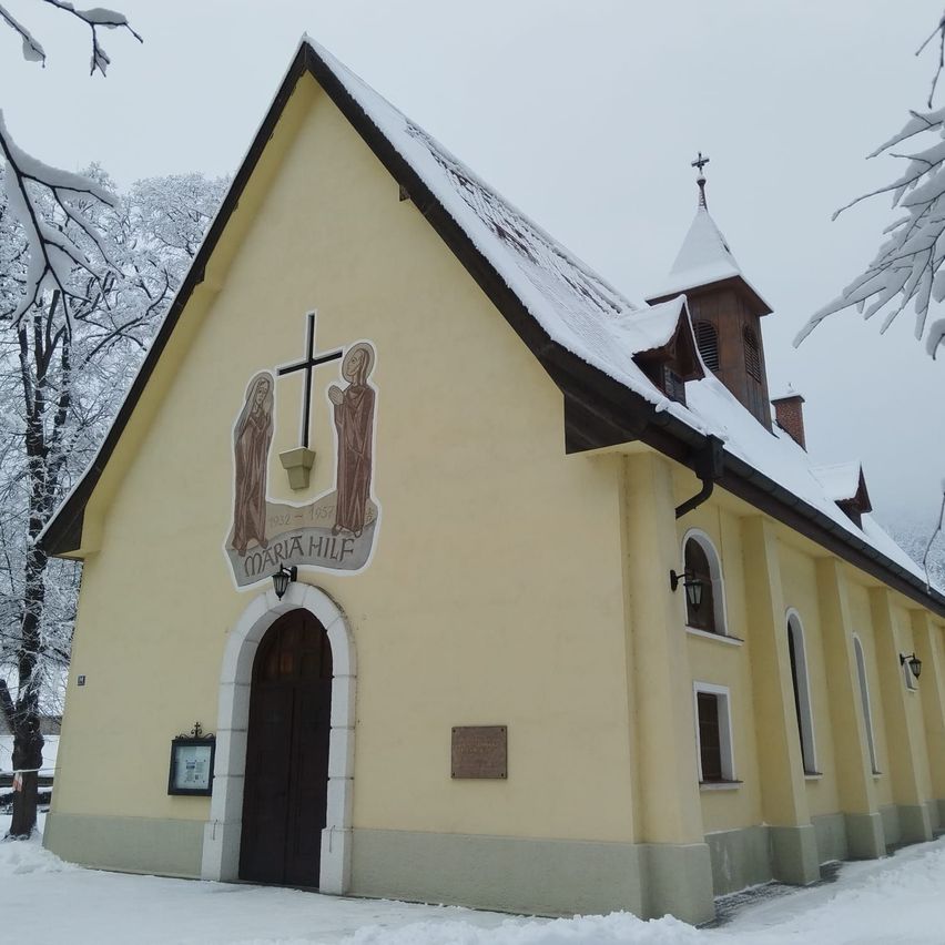 Eine gelbe Kirche mit einem Kreuz und Heiligenfiguren ist schneebedeckt. Der Name Maria Hilf steht über dem Eingang.