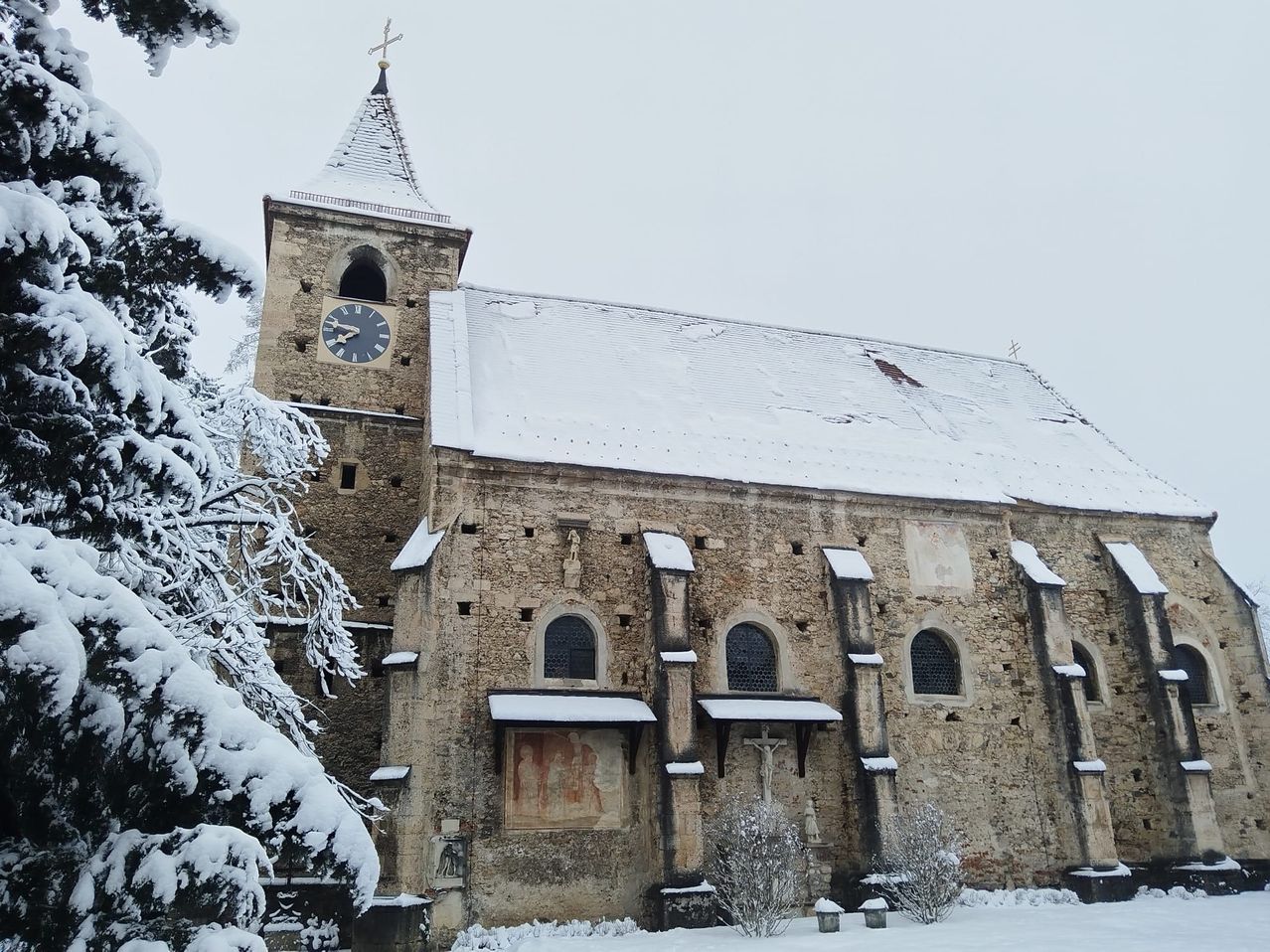Eine historische Kirche, die mit Schnee bedeckt ist. Sie hat einen Glockenturm mit einer Uhr, einem Kreuz oben und einem schneebedeckten Dach. Das Gebäude hat Bogenfenster und eine Wandmalerei.