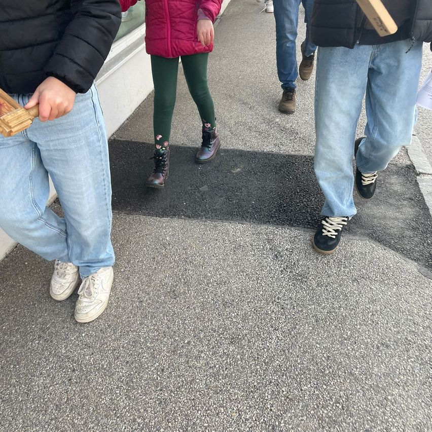 Vier Personen gehen auf einem gepflasterten Weg, jeder hält ein hölzerne Objekt. Der Weg hat eine frische schwarze Linie. Die Gruppe umfasst zwei Kinder in bunter Kleidung.