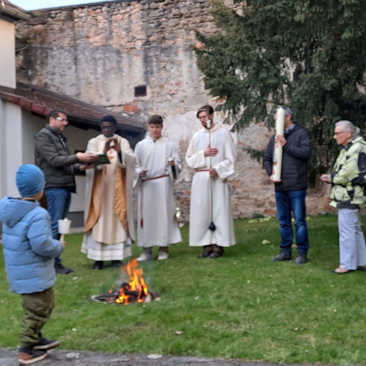 Eine Gruppe von Menschen in weißen Roben steht um ein Lagerfeuer. Ein Kind hält einen Becher. Zwei Männer halten Kerzen. Eine ältere Frau steht in der Nähe.