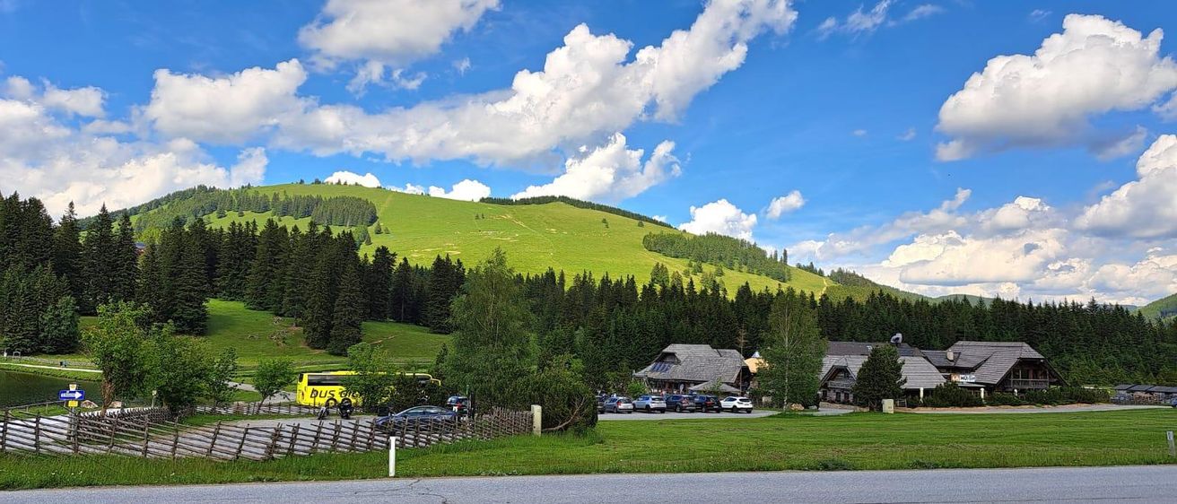 Ein landschaftlicher Blick auf einen Berg mit üppigem Grün, einem gelben Bus und mehreren Autos, die vor einer Berghütte geparkt sind. Der Himmel ist blau mit Wolken.