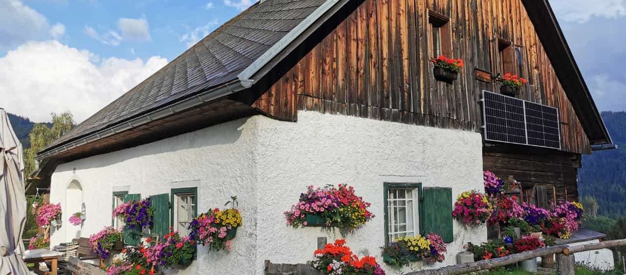 Ein charmantes rustikales Haus mit hölzerne Außenwand und weißer Basis, geschmückt mit bunten Blumen und einer Solaranlage auf dem Dach.
