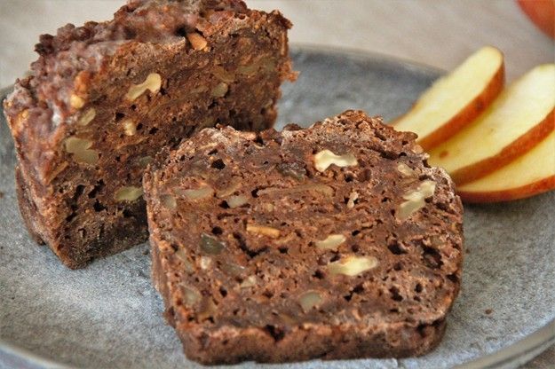 Ein Stück Schokoladen-Walnuss-Brot mit sichtbaren Nüssen auf einem grauen Teller mit Apfelscheiben.