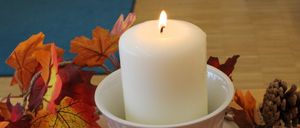 Eine brennende weiße Kerze steht in einem weißen Keramikbecher, umgeben von Herbstblättern und Tannenzapfen auf einer Holzoberfläche.