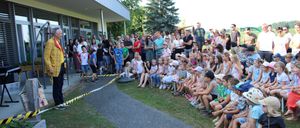 Eine Menschenmenge versammelt sich vor einem Gebäude, einige sitzen auf dem Rasen, während andere hinter einer Barriere stehen. Ein Mann in einem gelben Anzug spricht die Menge an.