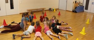 Eine Gruppe von Kindern liegt auf einem Holzboden mit mehreren Verkehrskegel verstreut. Eine erwachsene Frau kniet und lächelt die Kinder an.