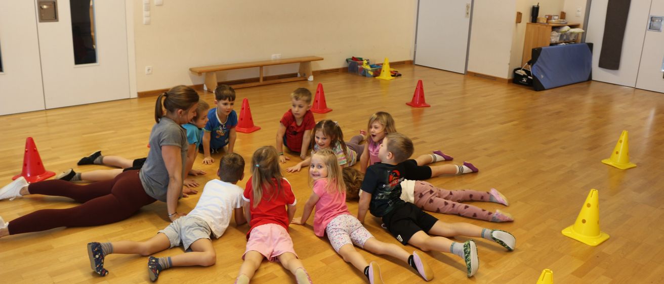 Eine Gruppe von Kindern liegt auf einem Holzboden mit mehreren Verkehrskegel verstreut. Eine erwachsene Frau kniet und lächelt die Kinder an.