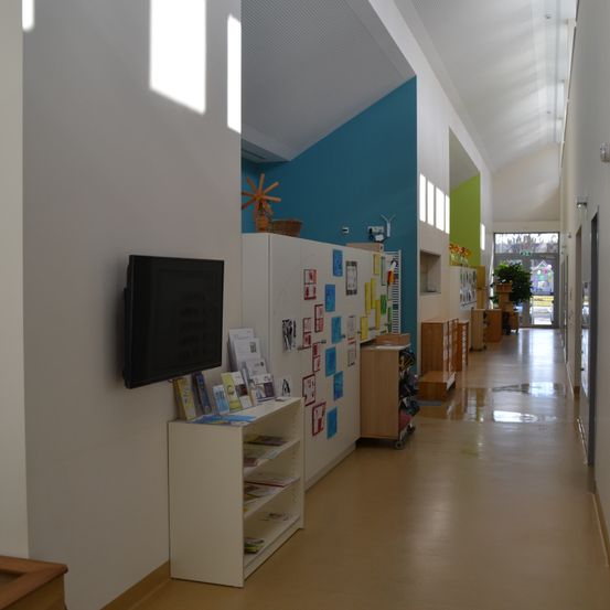 Ein leerer Flur mit weißen Wänden und einer blauen Akzentwand. Es gibt einen an der Wand montierten Fernseher, einen weißen Schrank mit Büchern und bunte Poster. Es gibt Schränke und eine Topfpflanze im Flur.