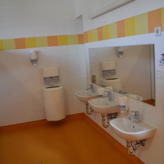 Ein Badezimmer mit weißen Wänden und orangefarbenen Fliesen. Es gibt drei Waschbecken, einen Spiegel und drei Papierhandtuchspender. Der Boden ist orange.