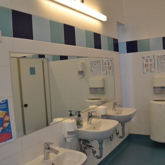 Ein Badezimmer mit drei Waschbecken und einem Spiegel, mit einer blau-weiß gefliesten Wand, einer Leuchtstofflampe und Handdesinfektionsspendern.