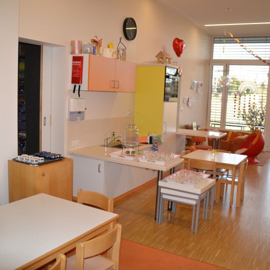 Eine Küche mit einem Waschbecken, Schränken und einer Arbeitsplatte mit verschiedenen Gegenständen. In der Nähe steht ein Tisch mit Stühlen, ein Schrank mit einer Mikrowelle und Dekorationen. Ein weiterer Tisch und Stühle befinden sich rechts. Im Hintergrund sind ein Fenster und eine Tür zu sehen.