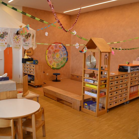 Ein Spielzimmer mit Holzmöbeln, einer kreisförmigen Dekoration an der Wand, einem Tisch mit Stühlen, einer Bank und verschiedenen Spielzeugen und Büchern auf Regalen.