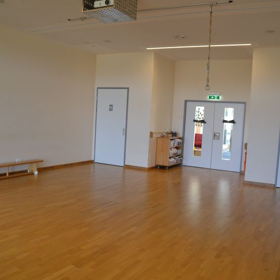 Ein leerer Raum mit polierten Holzböden, weißen Wänden und zwei Türen. Eine Bank und ein kleiner Bücherregal sind auf der linken Seite. Ein Projektor und ein hängender Seil sind an der Decke.
