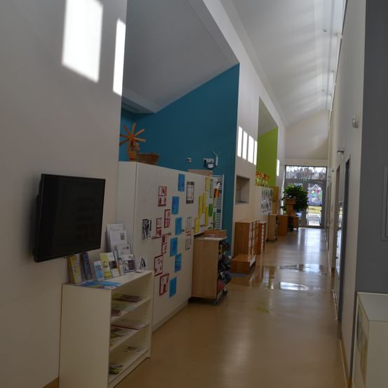 Ein leerer Flur mit weißer und blauer Wand, einem an der Wand montierten Fernseher und einem Regal mit Büchern und Papieren. Der Flur hat einen langen Korridor, Holzschränke und eine grüne Pflanze in der Ferne.