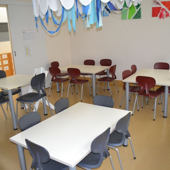 Ein leerer Raum mit mehreren weißen Tischen und burgunderroten Stühlen. Der Raum hat weiße Wände und eine Decke mit dekorativen Wimpelgirlanden.