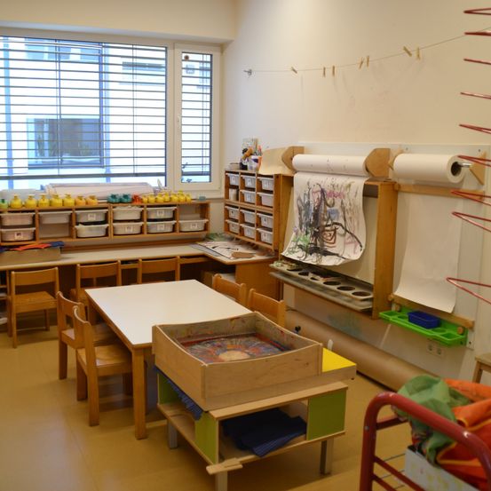 Ein heller, gut organisierter Kunstraum mit Tischen, Stühlen und Kunstmaterialien. Ein großes Fenster lässt natürliches Licht herein.