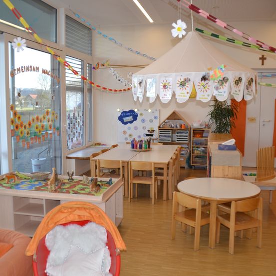 Ein Kindergartenraum mit einem Baldachin, bunten Dekorationen, Tischen, Stühlen und Spielzeug. Der Raum hat eine weiße Wand, eine Glastür und einen Blick auf einen Garten.