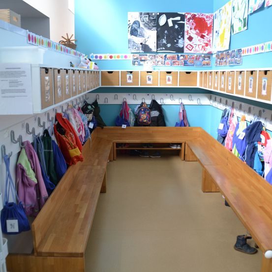 Ein Klassenzimmer mit blauen Wänden und Holzbänken. Verschiedene Aufbewahrungsboxen sind an der Wand montiert, mit mehreren Jacken und Taschen, die an Haken hängen. Kunstprojekte sind an der Wand ausgestellt.