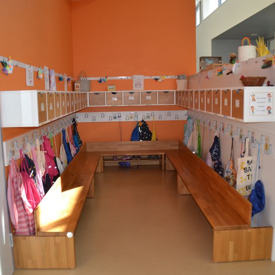 Ein heller, orange gestrichener Raum mit Holzbänken, Aufbewahrungsboxen und Haken zum Aufhängen von Kleidung. Verschiedene Taschen und Gegenstände sind verstreut.